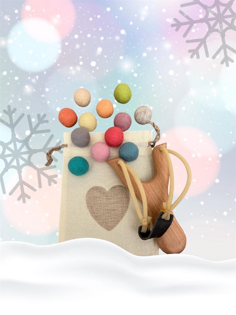 K&ouml;nnte beinhalten: Eine Holzschleuder mit einem schwarzen Lederbeutel und beigen Gummib&auml;ndern. Ein beiger Stoffbeutel mit Herzmuster ist mit bunten Filzkugeln gef&uuml;llt. Der Hintergrund hat ein Winterthema mit Schneeflocken und Bokeh.