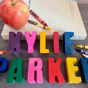 May include: A set of colorful, plastic letters spelling out the name "KYLIE PARKER". The letters are arranged in a line on a wooden surface. There are also crayons and an apple in the image.