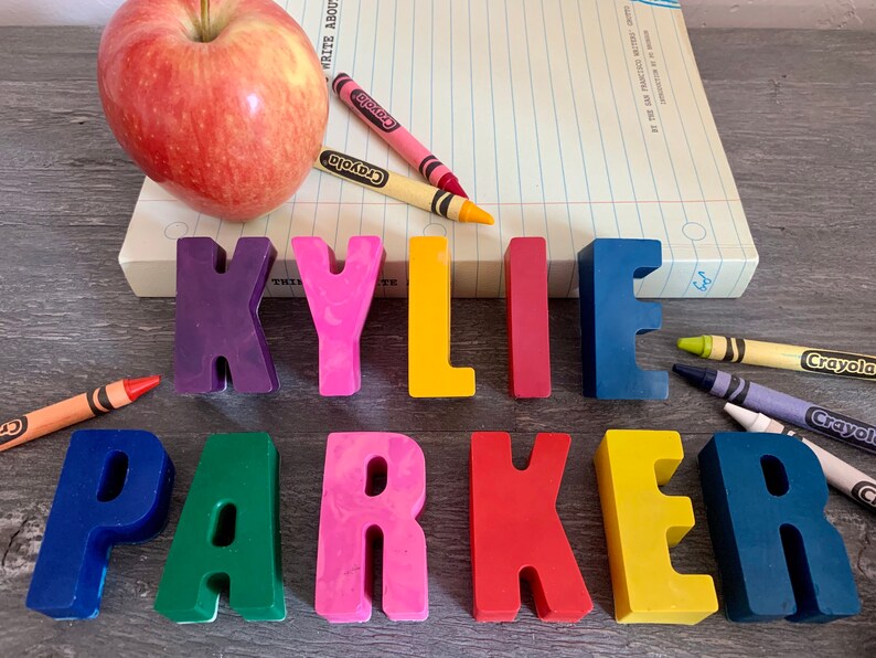 May include: A set of colorful, plastic letters spelling out the name "KYLIE PARKER". The letters are arranged in a line on a wooden surface. There are also crayons and an apple in the image.