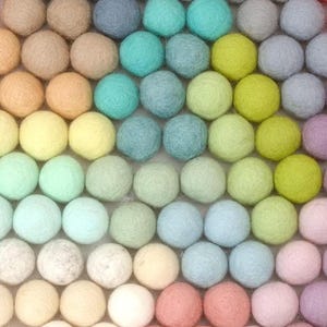 K&ouml;nnte beinhalten: Eine Anordnung farbenfroher Filzkugeln in Reihen. Die Kugeln zeigen einen Farbverlauf, darunter Schattierungen von Orange, Rosa, Blau, Gr&uuml;n, Gelb und Lila. Die Textur des Filzes ist sichtbar und erzeugt ein weiches, handwerkliches Aussehen.