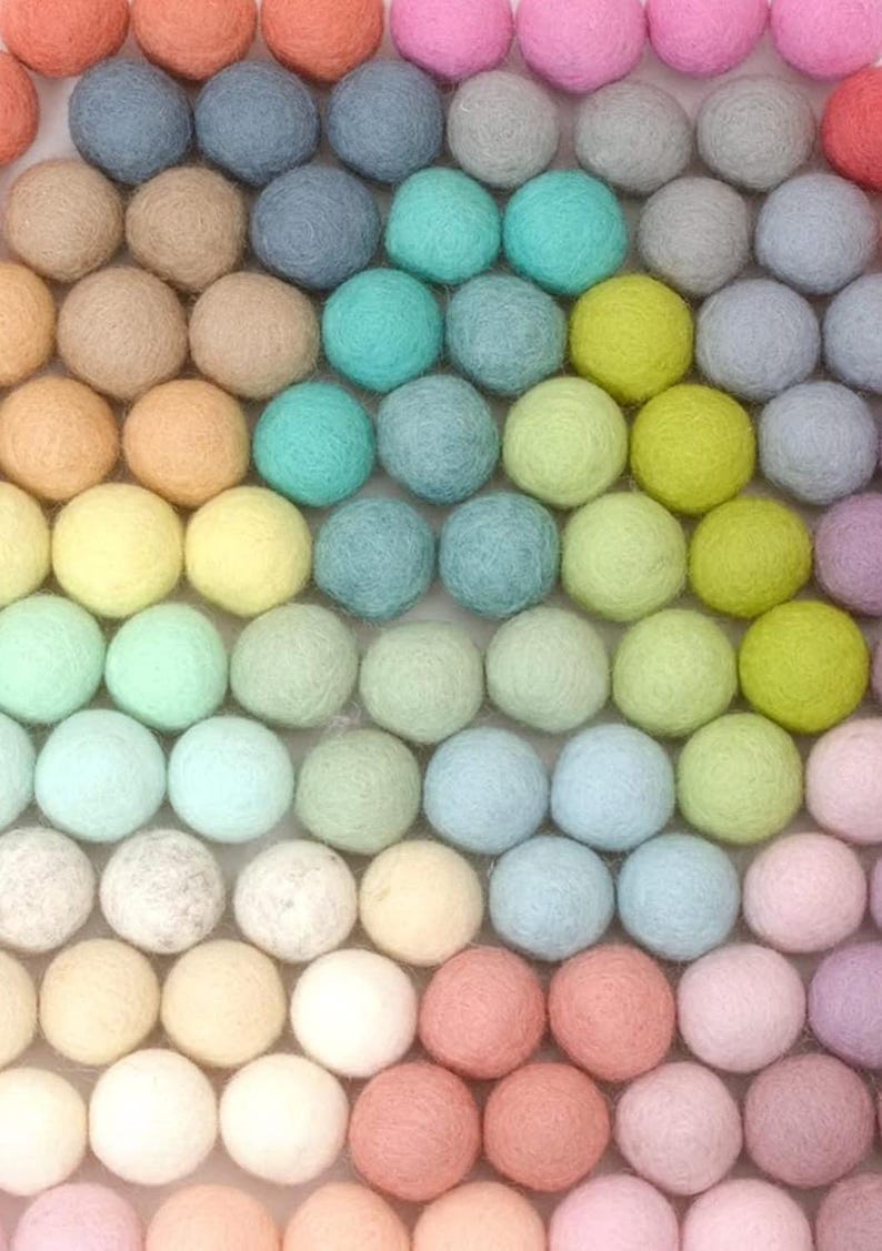 K&ouml;nnte beinhalten: Eine Anordnung farbenfroher Filzkugeln in Reihen. Die Kugeln zeigen einen Farbverlauf, darunter Schattierungen von Orange, Rosa, Blau, Gr&uuml;n, Gelb und Lila. Die Textur des Filzes ist sichtbar und erzeugt ein weiches, handwerkliches Aussehen.