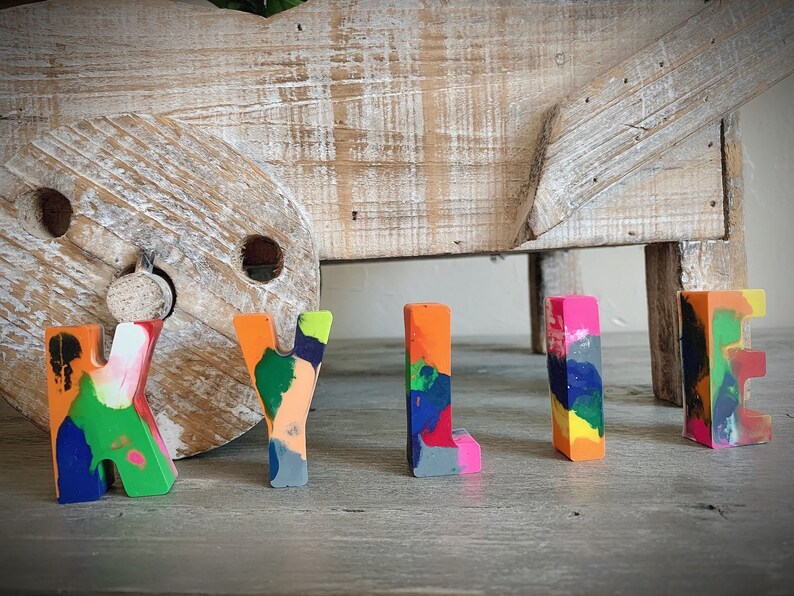 May include: The name "KYLIE" spelled out in colorful, multi-colored crayon letters. The letters are standing on a wooden surface.