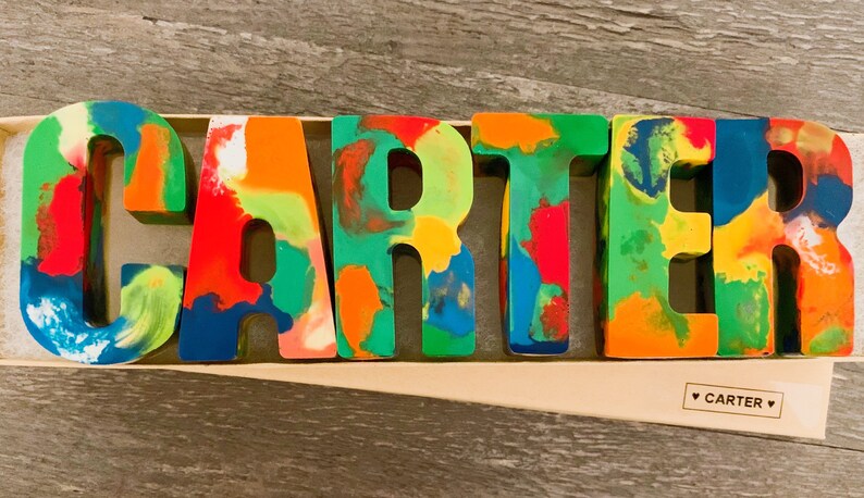 May include: Colorful, multi-colored, crayon letters spelling out the name "CARTER". The letters are in a box with a white label that says "CARTER" in a heart.