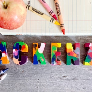 May include: The name Mackenzie spelled out in colorful crayon letters. The letters are made of melted crayons and are arranged on a wooden surface. There are also crayons and a notebook in the background.