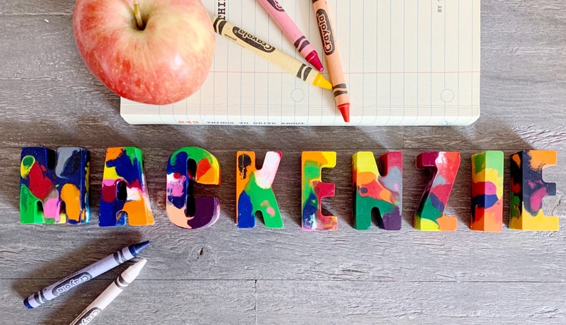 May include: The name Mackenzie spelled out in colorful crayon letters. The letters are made of melted crayons and are arranged on a wooden surface. There are also crayons and a notebook in the background.
