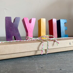 May include: A set of colorful wooden letters spelling out the name "KYLIE" in a rainbow color scheme. The letters are arranged in a row on a brown box with a white and red twine bow. The box is labeled "KYLIE".