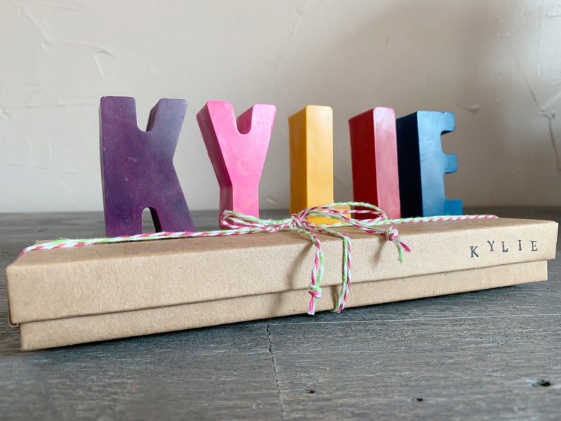 May include: A set of colorful wooden letters spelling out the name "KYLIE" in a rainbow color scheme. The letters are arranged in a row on a brown box with a white and red twine bow. The box is labeled "KYLIE".