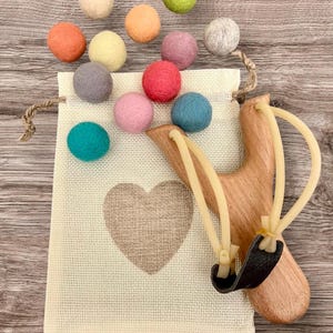 K&ouml;nnte beinhalten: Eine Holzschleuder mit schwarzem Lederbeutel und beigen Gummib&auml;ndern, neben einem beigen Kordelzugbeutel mit Herzmuster. Bunte Filzkugeln in Orange, Gelb, Rosa, Blau, Gr&uuml;n und Grau sind verstreut. Die Gegenst&auml;nde liegen auf einer Holzoberfl&auml;che.