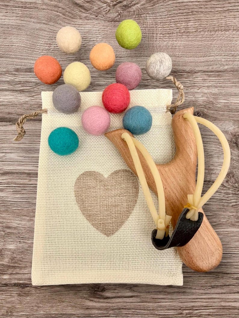 K&ouml;nnte beinhalten: Eine Holzschleuder mit schwarzem Lederbeutel und beigen Gummib&auml;ndern, neben einem beigen Kordelzugbeutel mit Herzmuster. Bunte Filzkugeln in Orange, Gelb, Rosa, Blau, Gr&uuml;n und Grau sind verstreut. Die Gegenst&auml;nde liegen auf einer Holzoberfl&auml;che.