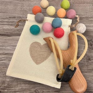 K&ouml;nnte beinhalten: Eine Holzschleuder mit einem schwarzen Beutel und einer Sammlung bunter Filzkugeln. Der Beutel ist beige mit einem Herzdesign. Der Text "Ready, Aim, Fun!" befindet sich am unteren Rand des Bildes.