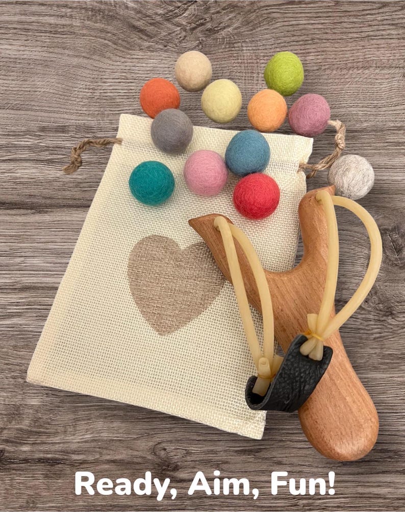 K&ouml;nnte beinhalten: Eine Holzschleuder mit einem schwarzen Beutel und einer Sammlung bunter Filzkugeln. Der Beutel ist beige mit einem Herzdesign. Der Text "Ready, Aim, Fun!" befindet sich am unteren Rand des Bildes.