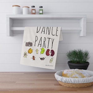 May include: A white kitchen towel with the words "DANCE PARTY" in black script. The towel features cartoon illustrations of dancing beans and a boombox. The towel is hanging on a white shelf above a wooden countertop.