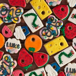 May include: A collection of colorful decorated cookies featuring the words "one" and "Luke" along with images of a caterpillar, a strawberry, a watermelon, and a piece of cheese.
