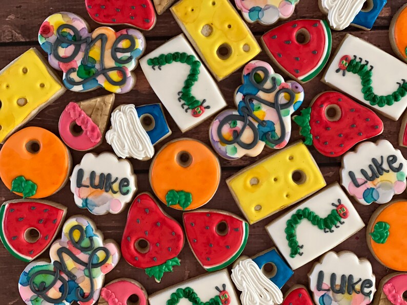 May include: A collection of colorful decorated cookies featuring the words "one" and "Luke" along with images of a caterpillar, a strawberry, a watermelon, and a piece of cheese.