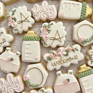 May include: A set of 14 decorated sugar cookies with various designs, including baby bottles, onesies, and floral arrangements. The cookies are decorated with pink, green, and gold icing. The cookies say "Baby" and "Welcome".