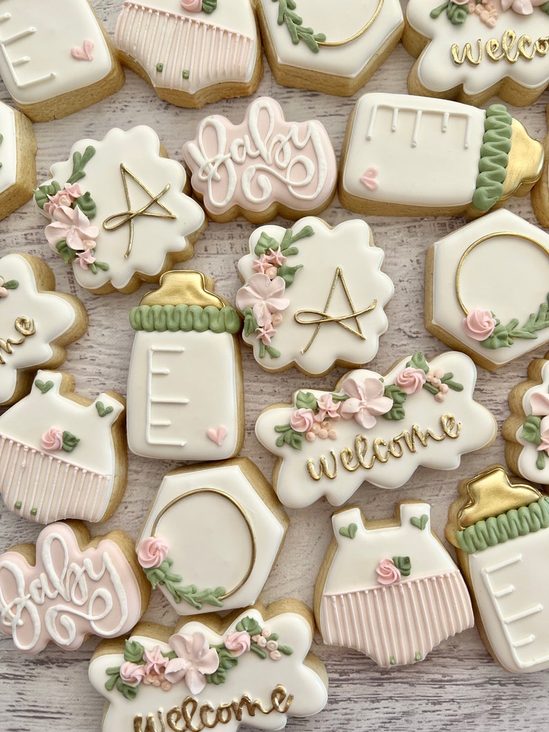 May include: A set of 14 decorated sugar cookies with various designs, including baby bottles, onesies, and floral arrangements. The cookies are decorated with pink, green, and gold icing. The cookies say "Baby" and "Welcome".