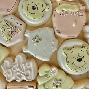 K&ouml;nnte beinhalten: Auswahl an dekorierten Zuckerkeksen mit Winnie-the-Pooh-Motiv. Die Kekse zeigen Pooh-B&auml;r-Gesichter, Honigt&ouml;pfe, Baby-Motive und das Wort "Baby" in Schreibschrift. Farben sind Gelb, Rosa, Wei&szlig; und Beige.