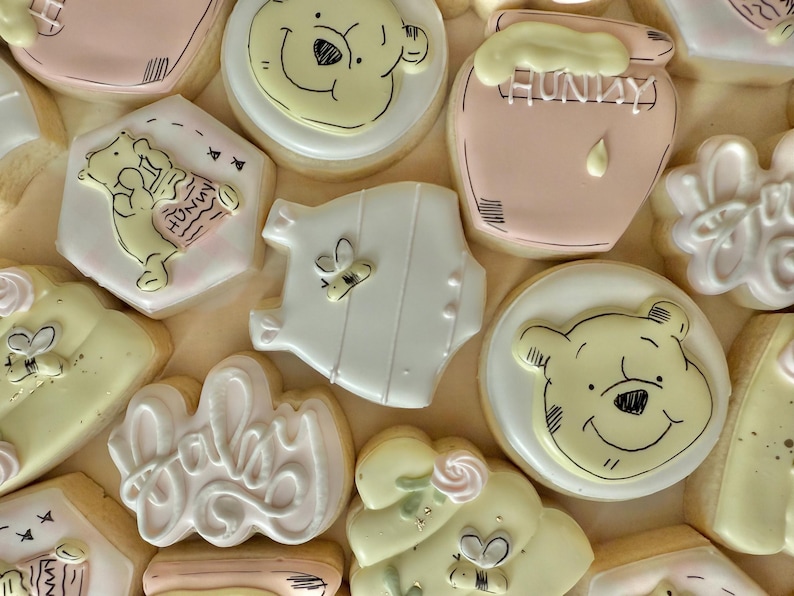 K&ouml;nnte beinhalten: Auswahl an dekorierten Zuckerkeksen mit Winnie-the-Pooh-Motiv. Die Kekse zeigen Pooh-B&auml;r-Gesichter, Honigt&ouml;pfe, Baby-Motive und das Wort "Baby" in Schreibschrift. Farben sind Gelb, Rosa, Wei&szlig; und Beige.