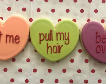 May include: Three heart-shaped cookies decorated with red icing and different messages: "eat me", "pull my hair", and "bend over".