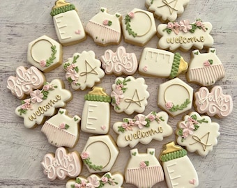 May include: A collection of 18 baby shower cookies decorated with pink and green icing. The cookies feature various designs, including baby bottles, onesies, floral wreaths, and the word "baby" written in cursive. Some cookies also have the word "welcome" written on them.
