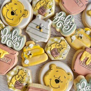 K&ouml;nnte beinhalten: Eine Auswahl an Keksen zum Thema Winnie Puuh. Die Kekse haben gelbe, wei&szlig;e und Pastellfarben. Einige sind in Form von Winnie Puuhs Gesicht, einem Baby-Strampler, einem Honigtopf und einer Honigwabe gehalten. Das Wort "Baby" steht auf einigen Keksen.
