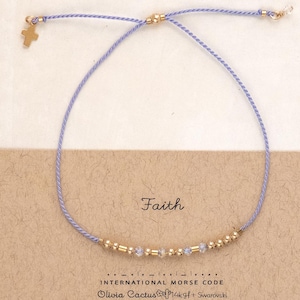 May include: A delicate bracelet with a light purple cord and gold-colored beads. A small gold cross charm hangs from the cord. The bracelet is displayed on a card that reads "Faith" in cursive.