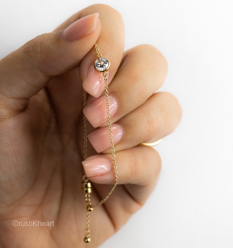 May include: A delicate gold chain bracelet featuring a single round, clear, bezel-set gemstone centered on the chain. The bracelet includes a sliding adjustable clasp mechanism with a small cylindrical component and a dangling gold bead at the end.