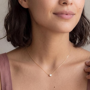May include: A delicate gold chain necklace featuring a single, round, clear gemstone set in a bezel setting. The necklace rests against the neck, showcasing a thin, polished gold metal chain that connects to the central, circular stone pendant.