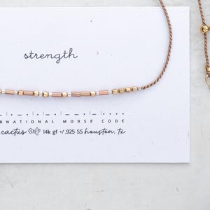 Può includere: Un delicato braccialetto con perline dorate e oro rosa, con la parola "strength" scritta sopra. Il braccialetto è su una carta bianca con codice Morse e il testo "olivia cactus 14k gf +.925 SS houston, tx."