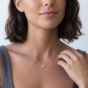 May include: A silver necklace featuring a single round, clear, faceted stone set in a bezel setting. The pendant is suspended from a delicate, thin silver chain that rests against the collarbone, creating a simple and refined appearance.