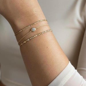 May include: A set of three gold-toned bracelets worn together. The top bracelet is a simple thin chain, the middle features a single round clear stone in a bezel setting, and the bottom bracelet consists of a delicate chain with small gold beads.