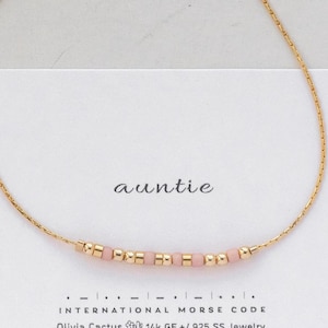 May include: A gold chain bracelet with pink beads spelling out "auntie" in international morse code. The bracelet is on a white card with the text "auntie" and "INTERNATIONAL MORSE CODE" printed on it.