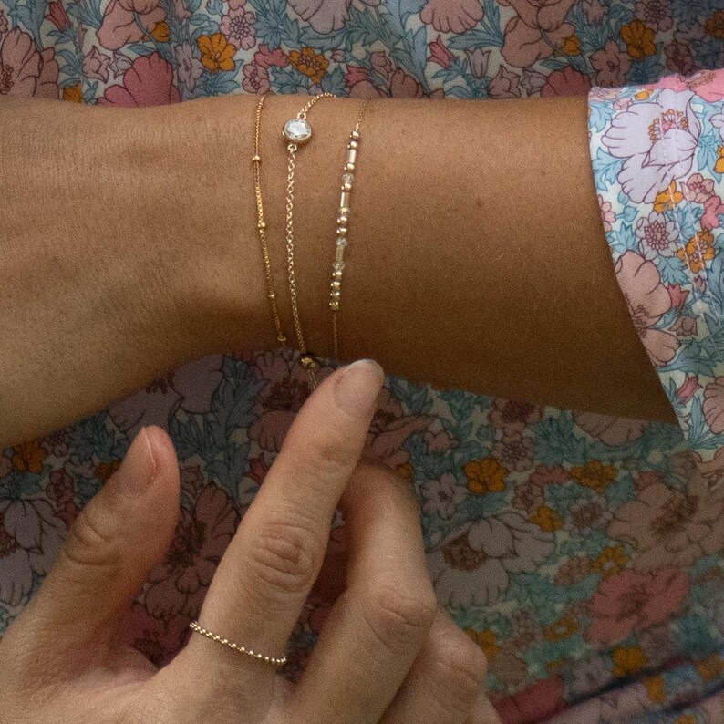 May include: Three delicate gold-toned chain bracelets worn together, including one with small beads, one with a single round clear stone, and one with a series of small metallic beads. A thin gold-toned beaded ring is worn on the index finger.