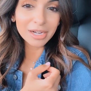 May include: A blue denim jacket featuring metal buttons, worn with a delicate gold chain necklace with a circular pendant, a second gold chain with a heart pendant, small stud earrings, a thin gold bracelet with a stone, and a silver-toned ring.