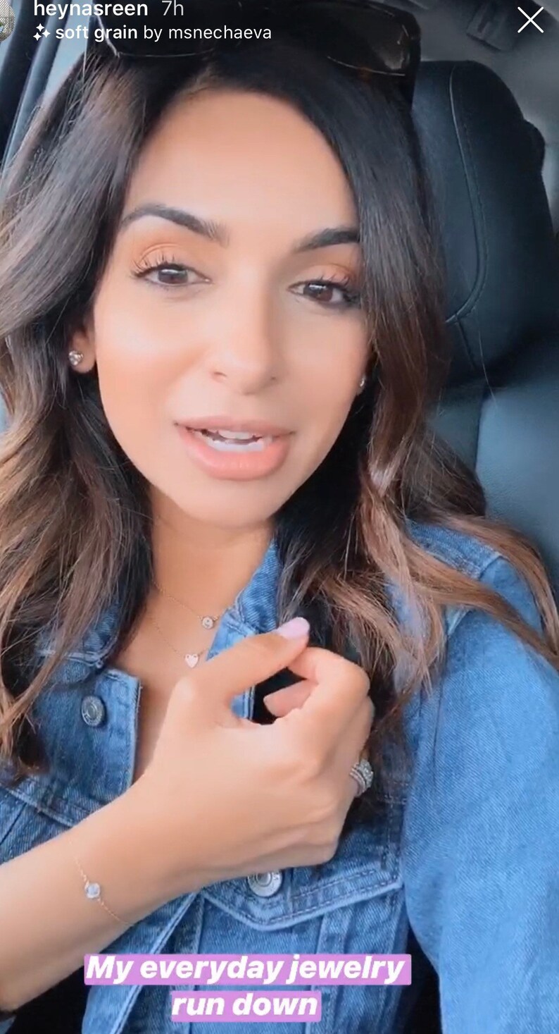 May include: A blue denim jacket featuring metal buttons, worn with a delicate gold chain necklace with a circular pendant, a second gold chain with a heart pendant, small stud earrings, a thin gold bracelet with a stone, and a silver-toned ring.