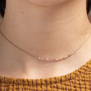 May include: A delicate gold chain necklace with small, evenly spaced beads. The necklace is worn by a person with a light complexion and brown hair.