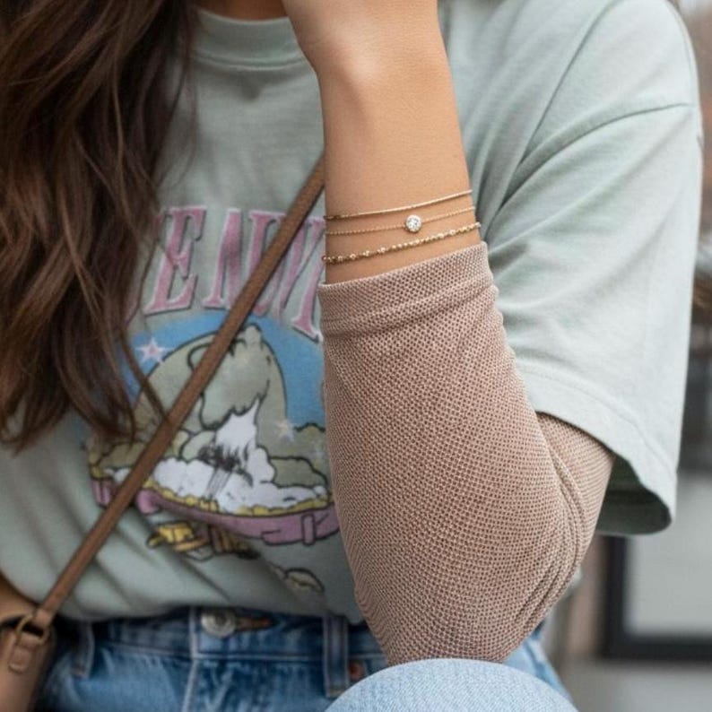 May include: Three delicate gold bracelets worn on the wrist. The top bracelet is a simple chain, the middle features a small round clear stone, and the bottom is a beaded chain. These are worn with a light green t-shirt and a tan long-sleeve shirt.
