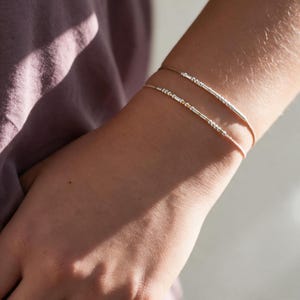 Könnte beinhalten: Zwei zarte Armbänder mit dünnen, hellen Kordeln, die mit kleinen, metallisch silberfarbenen Perlen und zylindrischen Abstandshaltern verziert sind, welche in einer an Morsecode erinnernden Abfolge angeordnet sind.