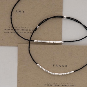 May include: Two black string bracelets with silver beads and bars spelling out the names "AMY" and "FRANK" in International Morse Code. The bracelets are on a brown background with the text "INTERNATIONAL MORSE CODE" printed below each name.
