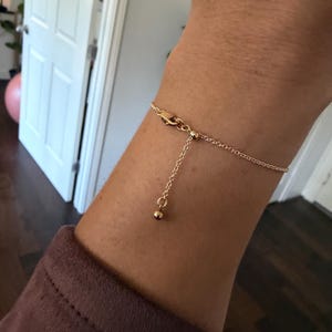 May include: A delicate gold-toned chain bracelet featuring a thin cable link design. The bracelet includes an adjustable sliding mechanism with a small round bead at the end of the dangling chain, secured by a lobster claw clasp closure.