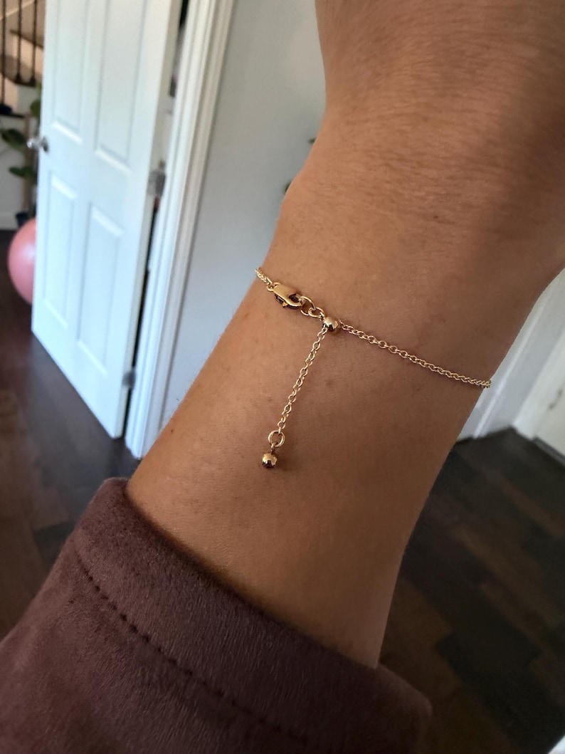 May include: A delicate gold-toned chain bracelet featuring a thin cable link design. The bracelet includes an adjustable sliding mechanism with a small round bead at the end of the dangling chain, secured by a lobster claw clasp closure.
