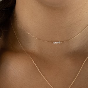 May include: A delicate gold chain necklace featuring three small, round white pearls centered on the strand. The necklace is worn as a choker, accompanied by a second, longer gold chain necklace that hangs below the primary pearl-accented piece.