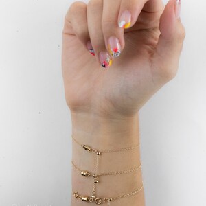 May include: Three gold-toned chain bracelets featuring small cylindrical magnetic clasps and delicate link chains. The bracelets are connected by a vertical chain segment with a small bead detail, creating a layered appearance on the wrist.