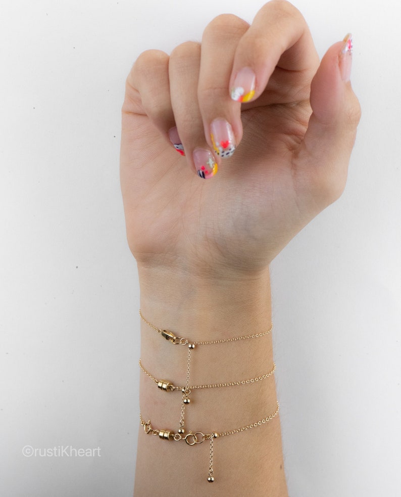 May include: Three gold-toned chain bracelets featuring small cylindrical magnetic clasps and delicate link chains. The bracelets are connected by a vertical chain segment with a small bead detail, creating a layered appearance on the wrist.