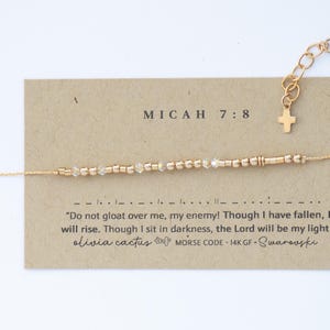 May include: A gold-coloured Morse code bracelet with clear crystal beads and a small cross charm. The bracelet is displayed on a tan card with the text "MICAH 7:8" and a quote. The brand name "olivia cactus" is also visible.