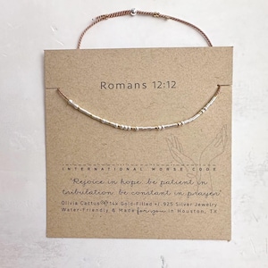 May include: A silver and gold beaded bracelet with a brown cord. The bracelet has the text "Romans 12:12" and the quote "Rejoice in hope, be patient in tribulation. be constant in prayer" on a brown card.