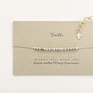 May include: A gold-colored bracelet with beads spelling out "Faith" in Morse code. The bracelet is on a tan card with the word "Faith" written in cursive. The card also has the text "International Morse Code" and "Olivia Cactus".