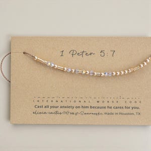 May include: A gold-colored beaded bracelet with clear crystal accents is displayed on a tan card. The card features the text "1 Peter 5:7" and a Morse code message. The text "Cast all your anxiety on him because he cares for you" is also visible.