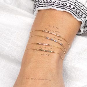 May include: Four thin gold chain bracelets featuring small coloured beads arranged in Morse code patterns. The bracelets are labelled with the words auntie, tribe, and nana, accompanied by their corresponding Morse code dots and dashes.
