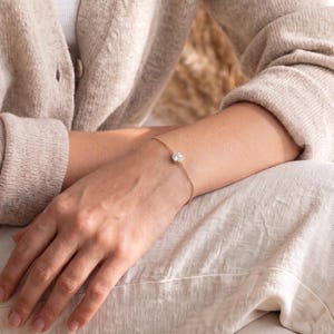 May include: A delicate gold chain bracelet featuring a single, round, clear faceted stone set in a simple gold bezel. The bracelet rests on a wrist, showcasing a minimalist design with a thin, polished metal chain and a secure, centered gemstone.